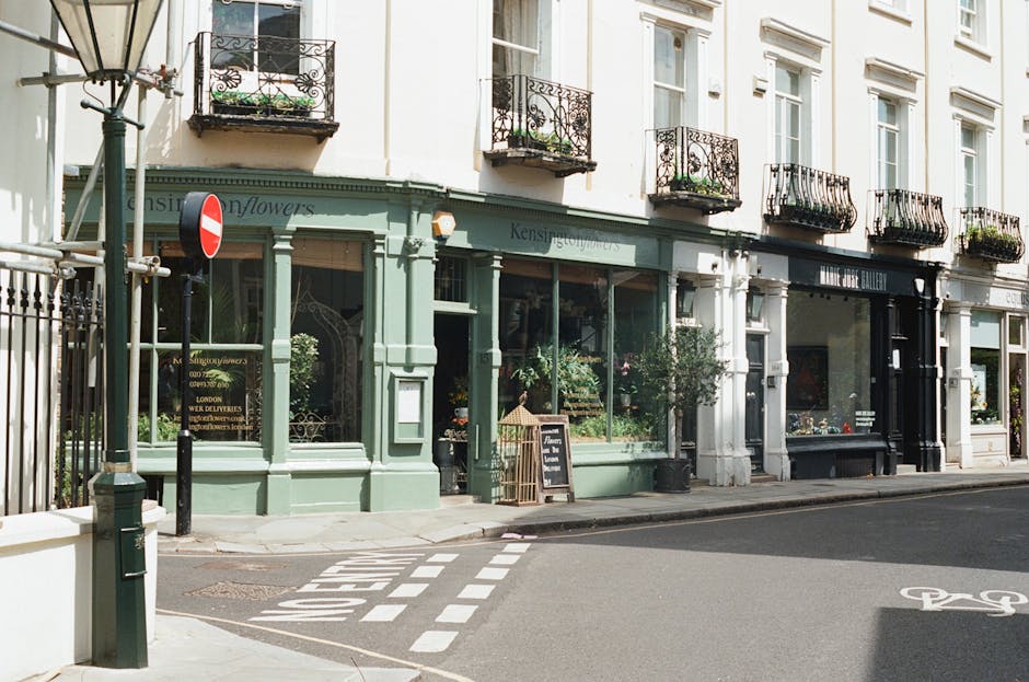A floral storefront on a city street corner, featuring large display windows filled with fresh flowers in various shades of pink, white, and green. The shop's exterior is painted in pastel green with 