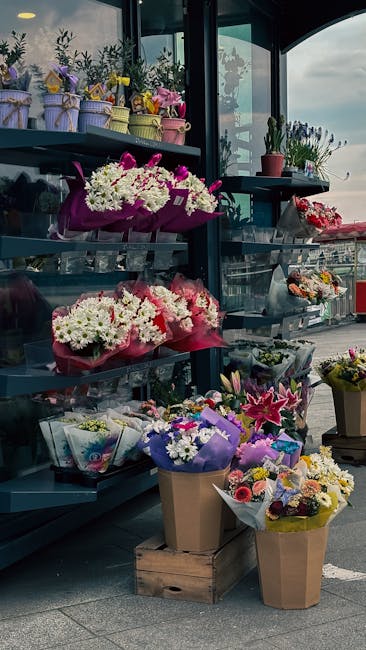 A display of fresh floral arrangements and bouquets situated outdoors on dark blue tiered shelves. The flowers include white daisies, pink lilies, purple irises, and multicolored roses, all wrapped in