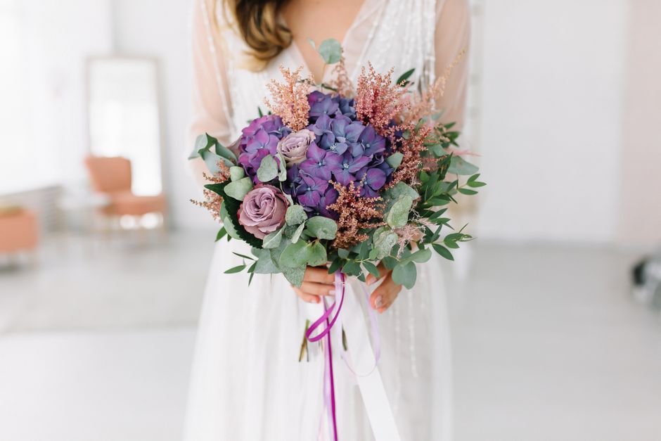 A person wearing a white dress holds a round floral bouquet featuring purple hydrangeas, soft lavender roses, and pink astilbe, complemented by green eucalyptus leaves. The bouquet is wrapped with lig