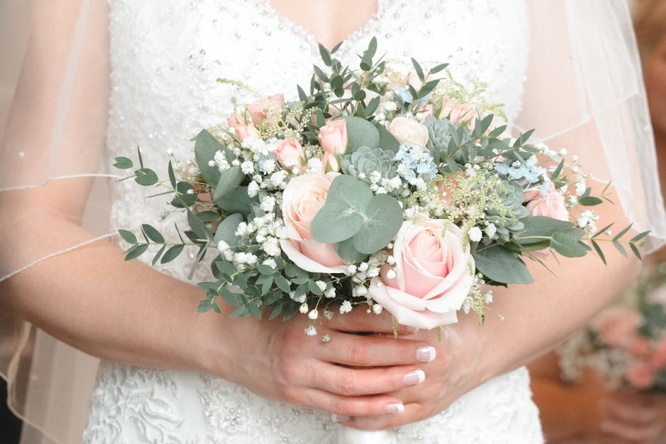 A bride in a white wedding gown holds a floral bouquet composed of pale pink roses, white baby's breath, and green eucalyptus leaves, arranged in a rounded, lush style. The bouquet is wrapped with a d