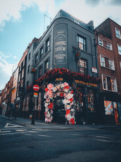 A storefront on Old Compton Street in Soho decorated with a large, vibrant floral arch made of pink, white, and red roses and peonies, arranged around the entrance. Above the arch, there is a hanging