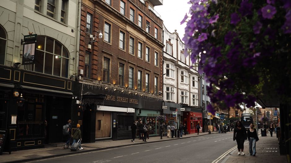 A street scene in Soho on Old Compton Street featuring a variety of brick and white-painted buildings, including the George Street Hotel with a black facade and gold lettering. The street is lively wi
